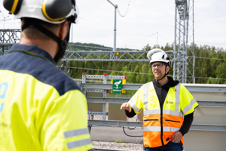 Två personer i gula varselvästar, med hjälmar på huvudet, står i området för en kraftstation.